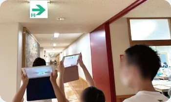 校舎の廊下で、タブレットを掲げて非常口の案内を確かめている生徒たち
