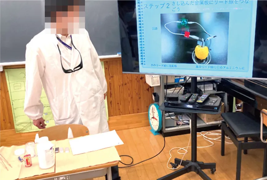 白衣を着た先生が、金属板にリード線をつなぐ手順を大型モニターに映して説明している