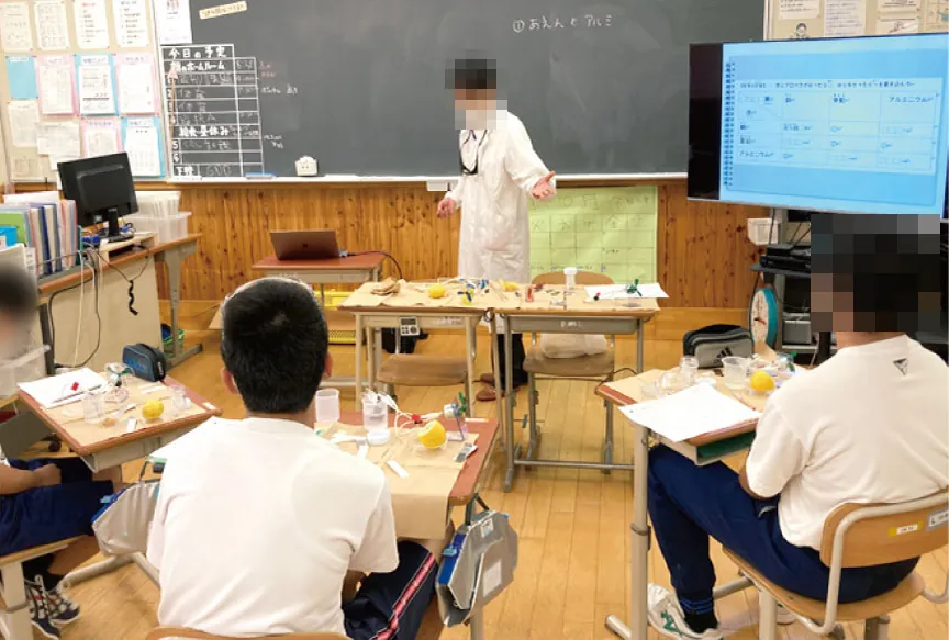 教室の前で説明する先生と、レモンや実験器具が置かれた机に向かう生徒たち