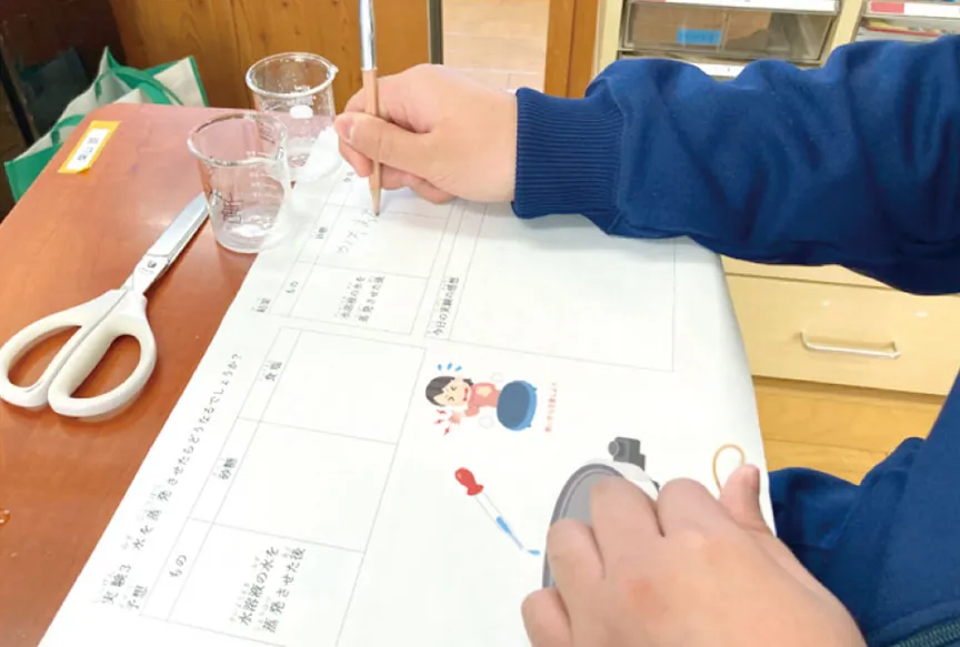 ワークシートに実験の結果を書き込んでいる手元。机にはビーカーとはさみがある