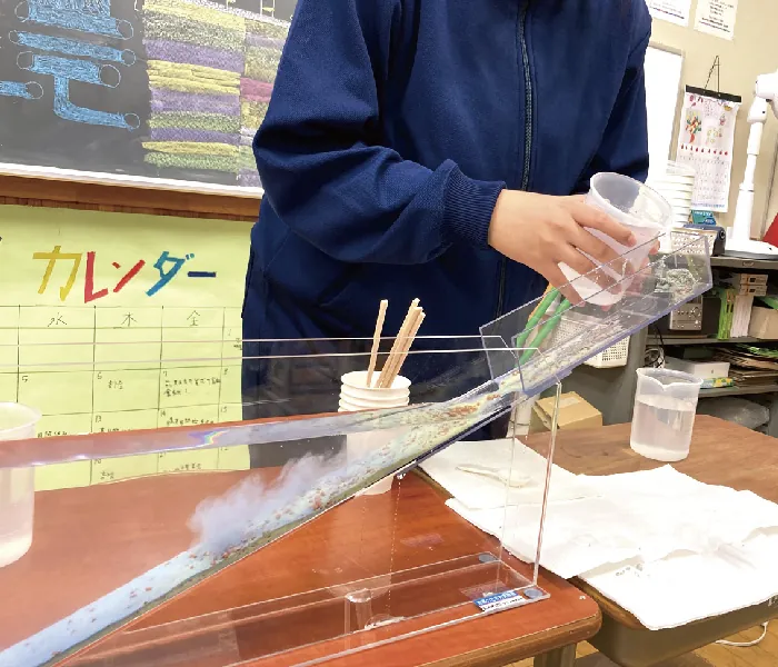 粒の大きさが異なる砂を水で流して積もり方を調べる実験装置と、水を注ぐ生徒